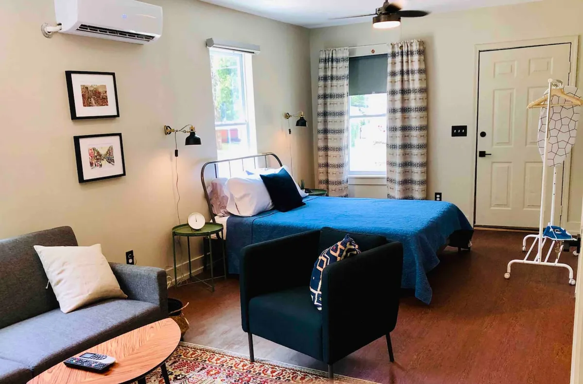 A bright bedroom with a bed dressed in blue bedding, a sofa adorned with decorative pillows, framed artwork, and a window letting in plenty of natural light. The space feels welcoming, perfect for relaxing, reading, or sharing moments with family.