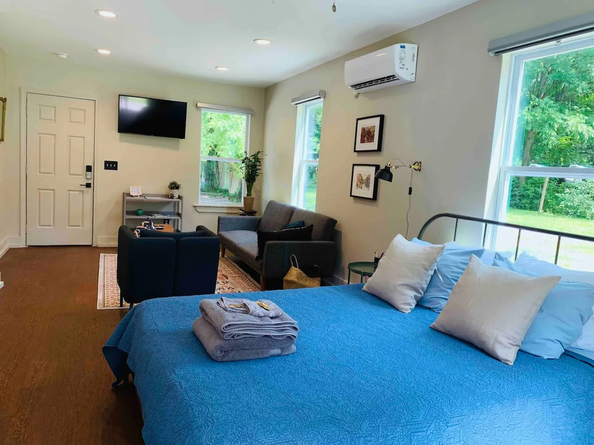 A spacious bedroom featuring a bed with blue bedding, a cozy sofa seating area, a television, and large windows offering a lush view of greenery outside. The scene is well-lit and exudes a comfortable, homey atmosphere.