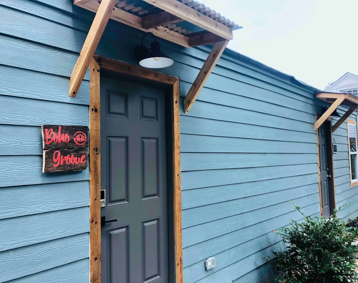 This image showcases the exterior of the duplex, featuring blue wooden siding, a dark gray door, a wooden awning, and a decorative sign by the entrance. The scene gives a friendly, welcoming impression, ideal for greeting families returning from outings in Overton Park.