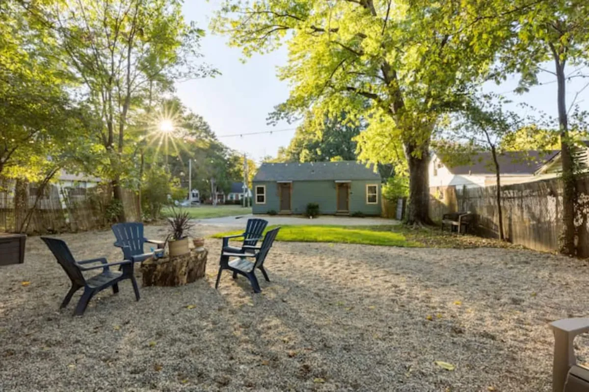 An outdoor scene with a fire pit surrounded by Adirondack chairs, set against a green house and shaded by mature trees. The space exudes a cozy, social atmosphere ideal for winding down in the evenings or enjoying the outdoors with loved ones. The trees provide ample shade, making the backyard inviting throughout the day.