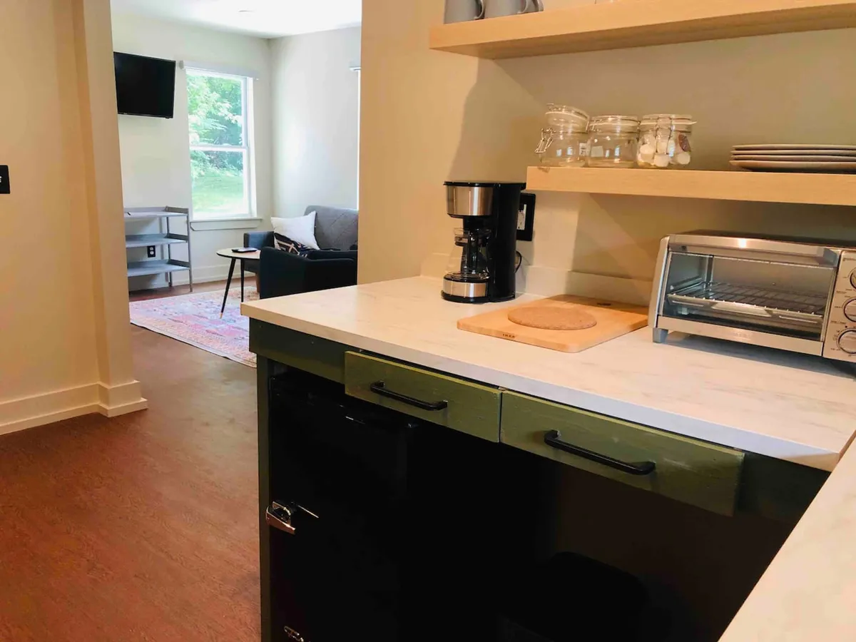 The image features a cozy kitchen area with a coffee maker and toaster on the counter, shelves lined with jars, and a sleek black refrigerator. The setting is styled for comfort and casual interaction, evoking a welcoming mood for morning rituals or preparing a light meal.