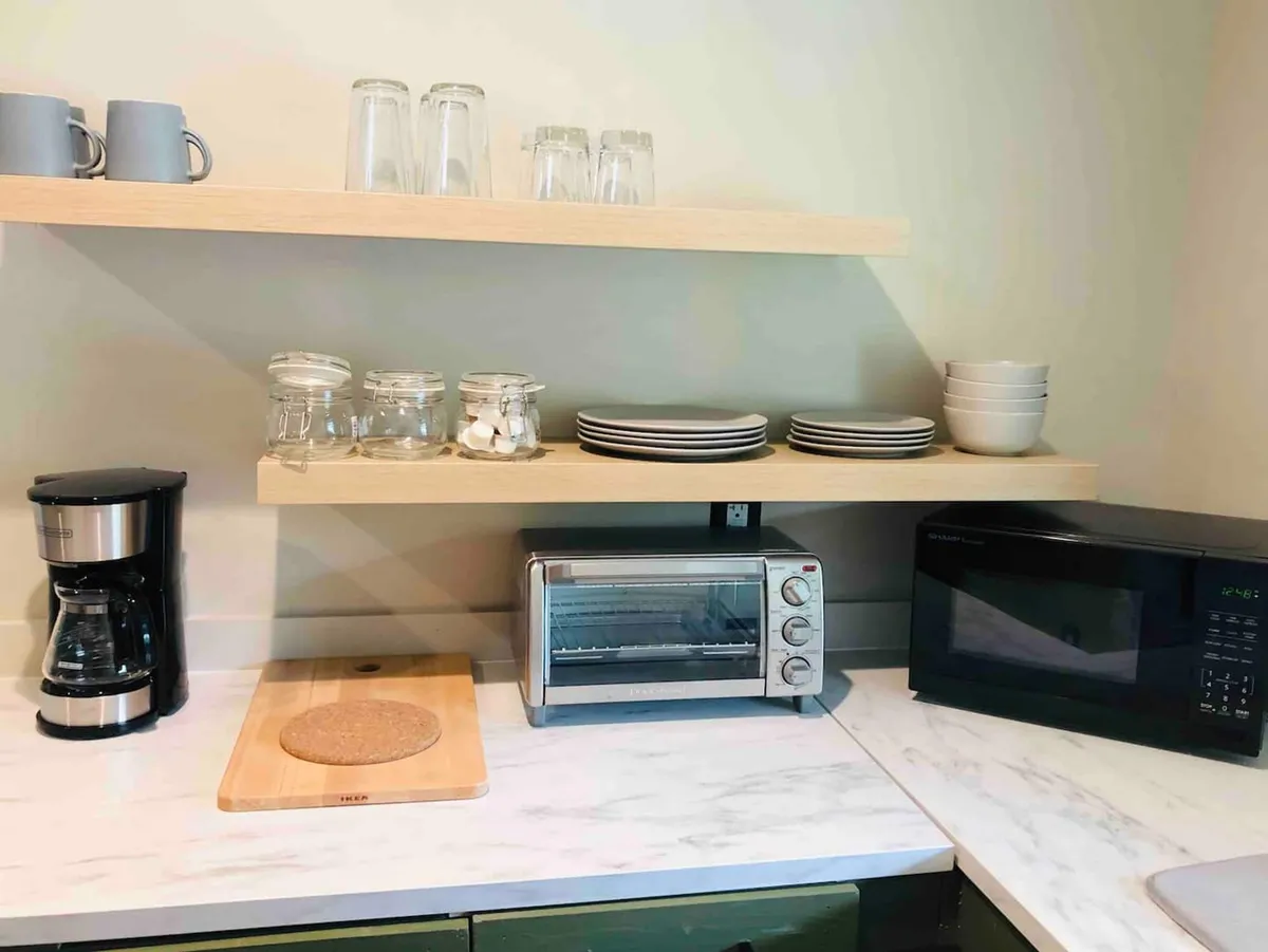 A functional kitchen corner showcasing a coffee maker, toaster oven, microwave, and shelves filled with glassware. The scene is homey and organized, emphasizing the convenience of meal prep and breakfast routines.