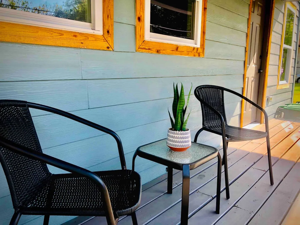 A pair of chairs and a small table sit against a blue wall with potted plants and framed windows nearby. The scene suggests an inviting nook for enjoying coffee, reading, or evening chats outdoors in a relaxed, homey environment.