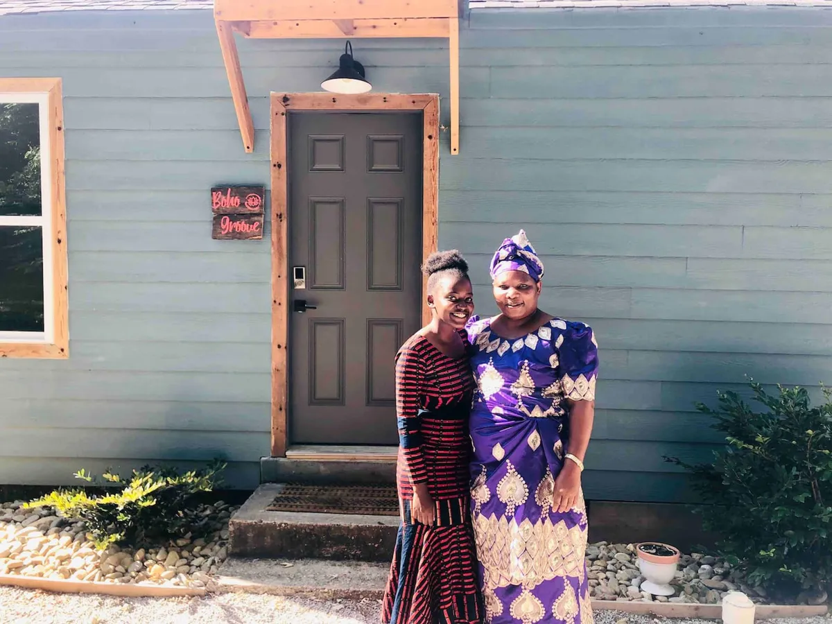 Two women in traditional clothing stand at the front door of the home, with a 'Bobo' and 'Private' sign visible. The mood is joyful and sociable, hinting at cultural exchange and outdoor gatherings.