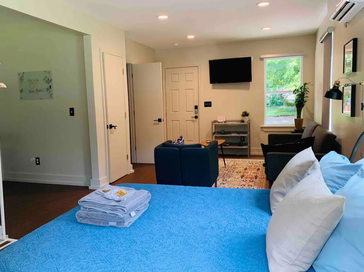 A well-lit room featuring a bed with a blue cover and a comfortable seating area with a couch and chairs. A television is mounted on the wall, and natural light streams in through a window, creating a welcoming space for relaxation and togetherness.