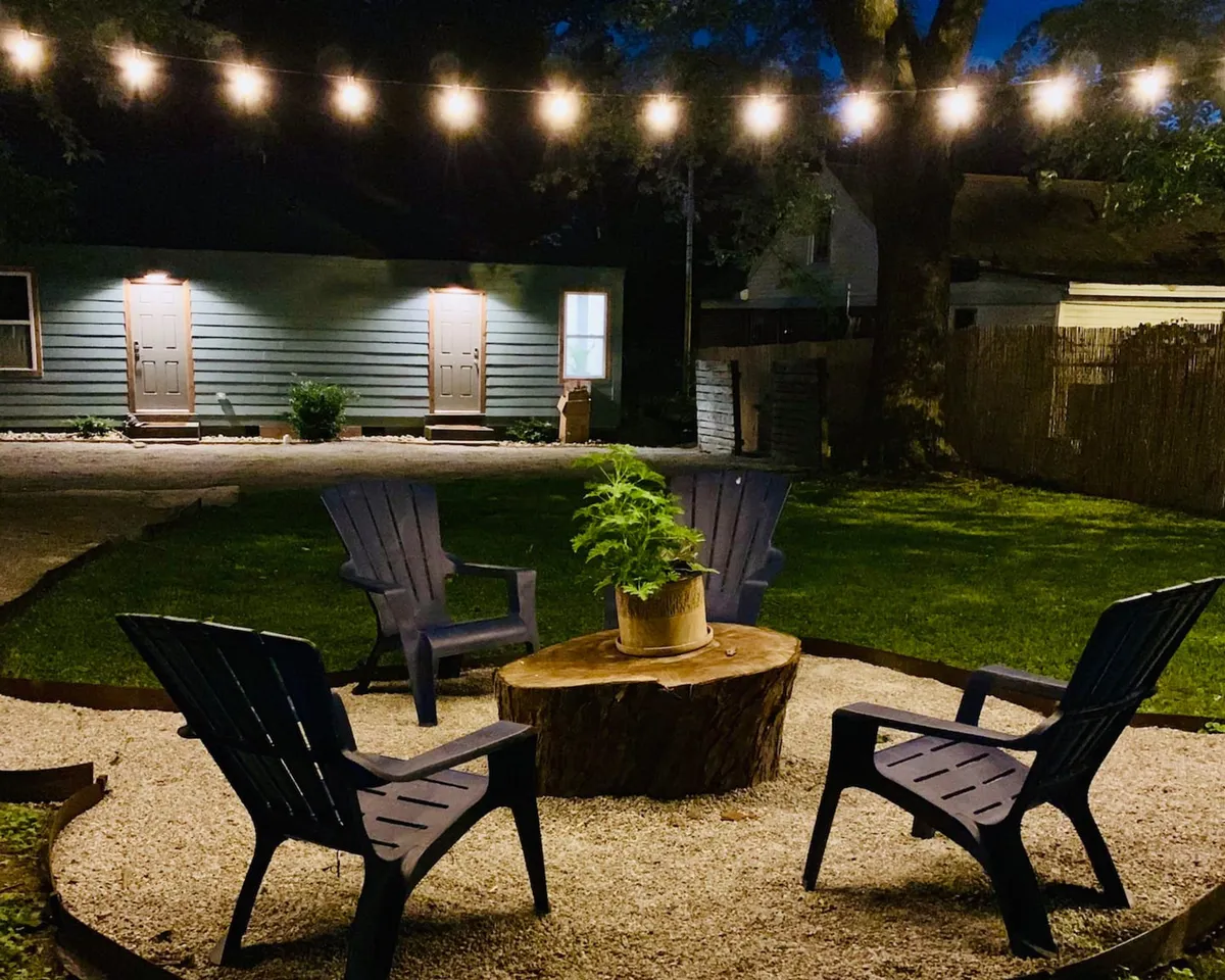 The image shows a cozy backyard setting at dusk, featuring a glowing fire pit surrounded by adirondack chairs, twinkling string lights overhead, and lush greenery creating a private, inviting atmosphere. The scene radiates warmth and comfort, perfect for evening gatherings or quiet moments under the stars.